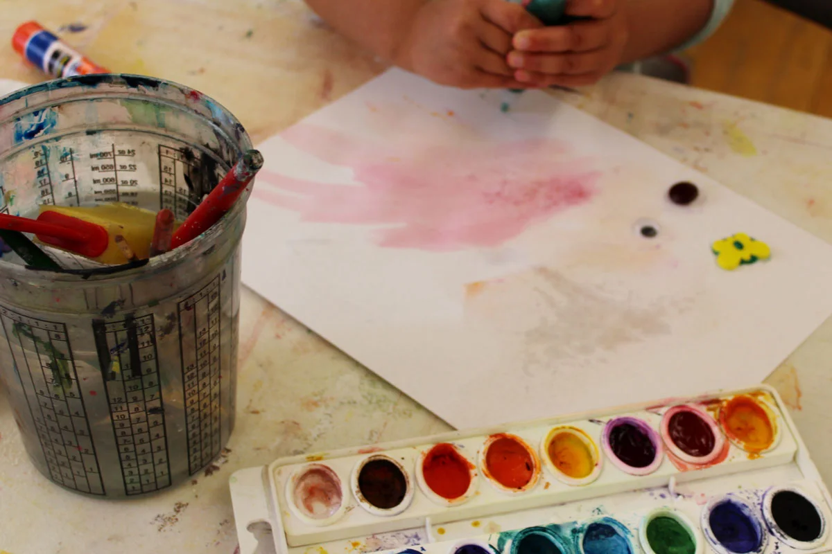 Child painting with watercolors beside a palette and paintbrush container.