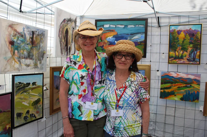 Two people standing in an art booth surrounded by colorful paintings.