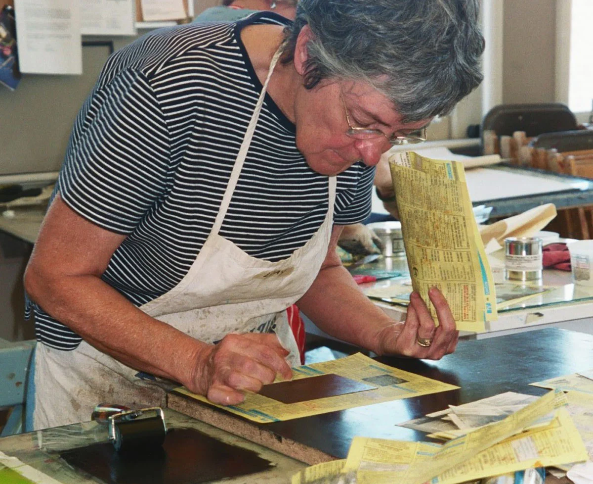 Person in a striped shirt and apron engaging in printmaking activities at a workbench.