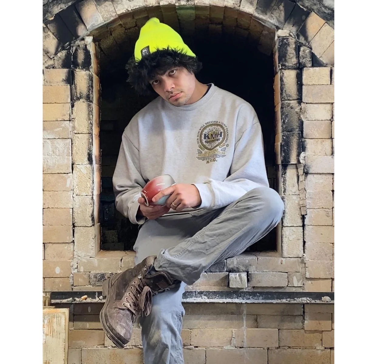 Person in a yellow beanie and grey sweatshirt sitting in a brick kiln opening, holding a cup.