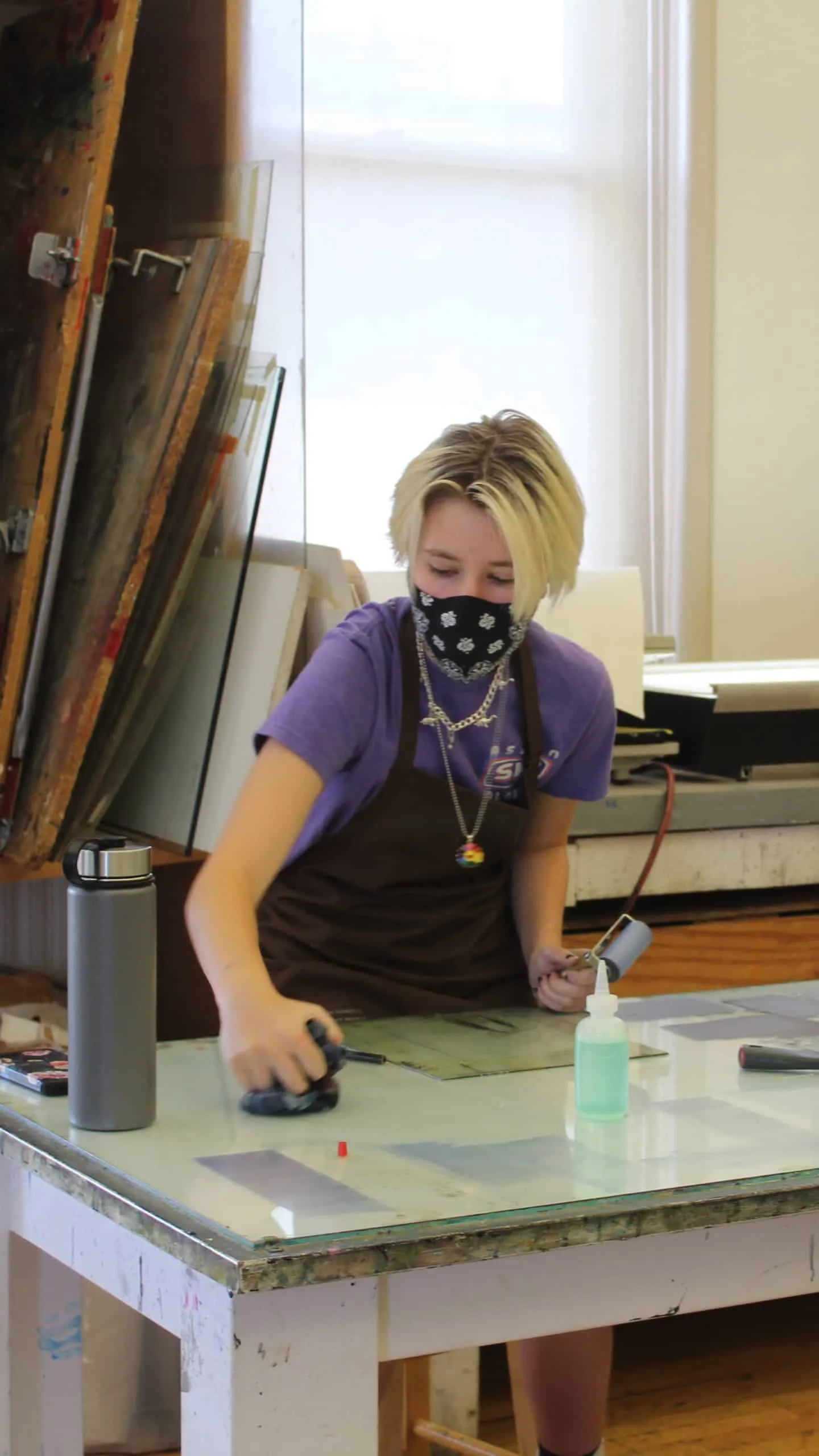 Person in apron working with printmaking tools on a glass surface in an art studio.