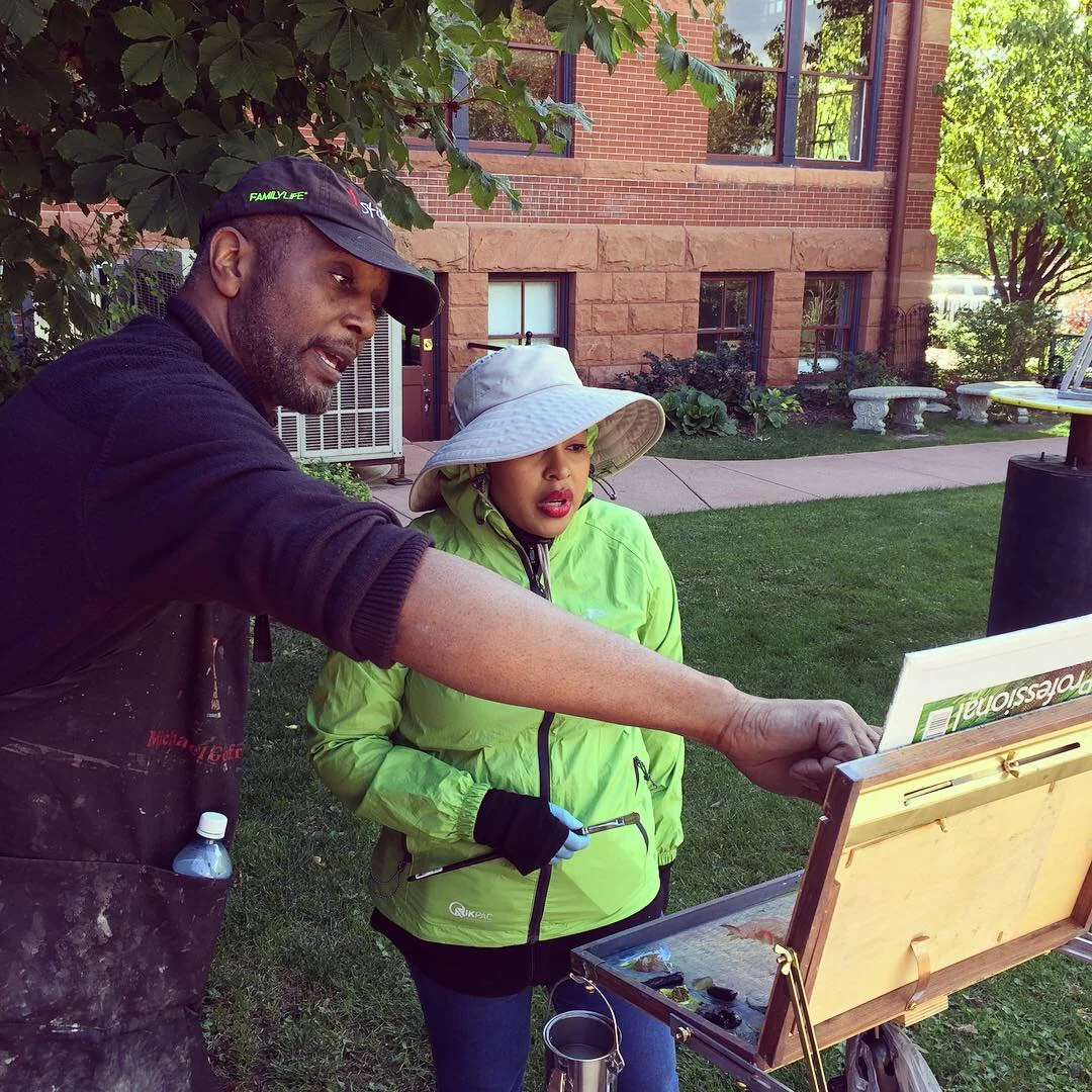 Two people painting outdoors with an easel and garden in the background.