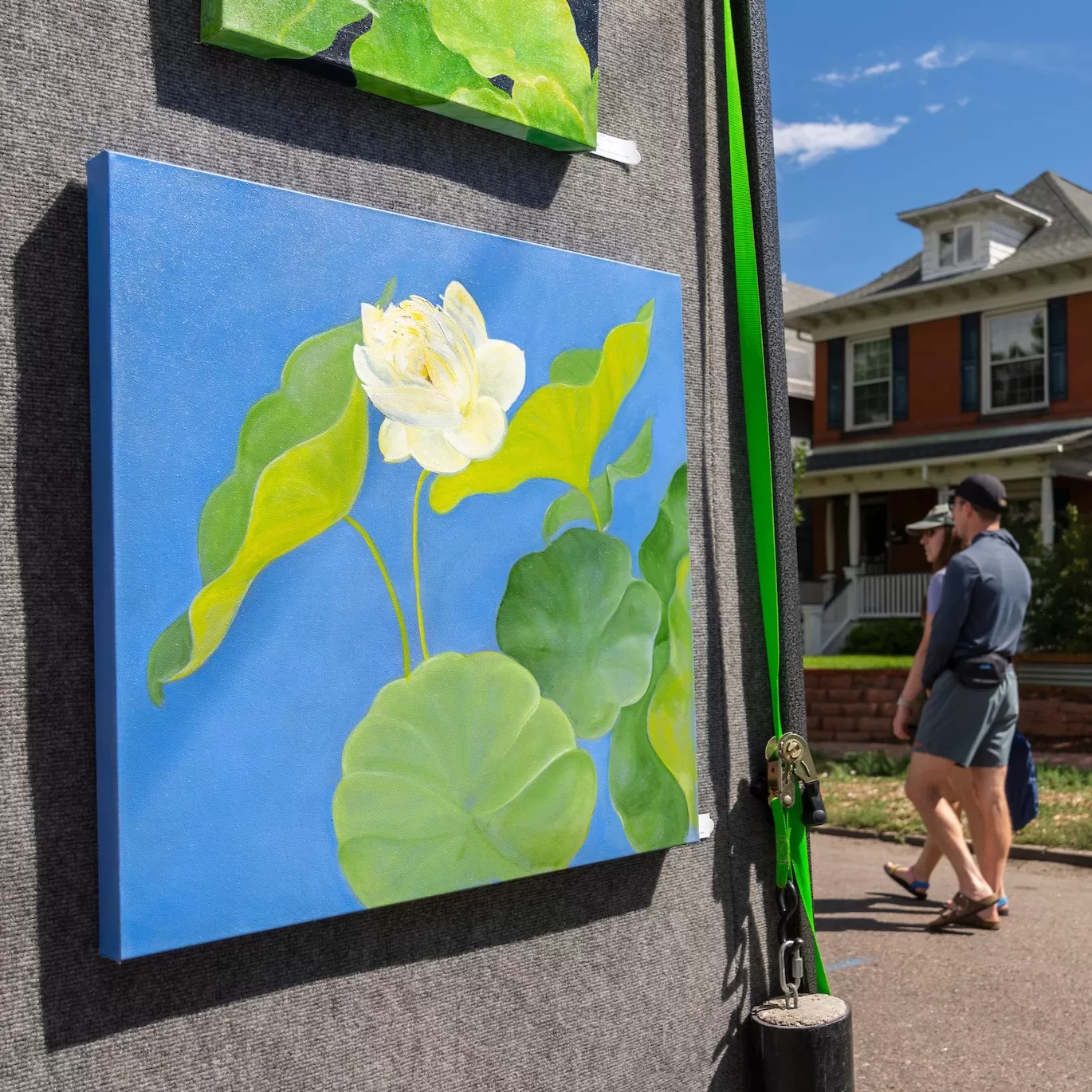 Painting with a blue background and a white flower with green leaves hung on the outside of an artists booth tent.