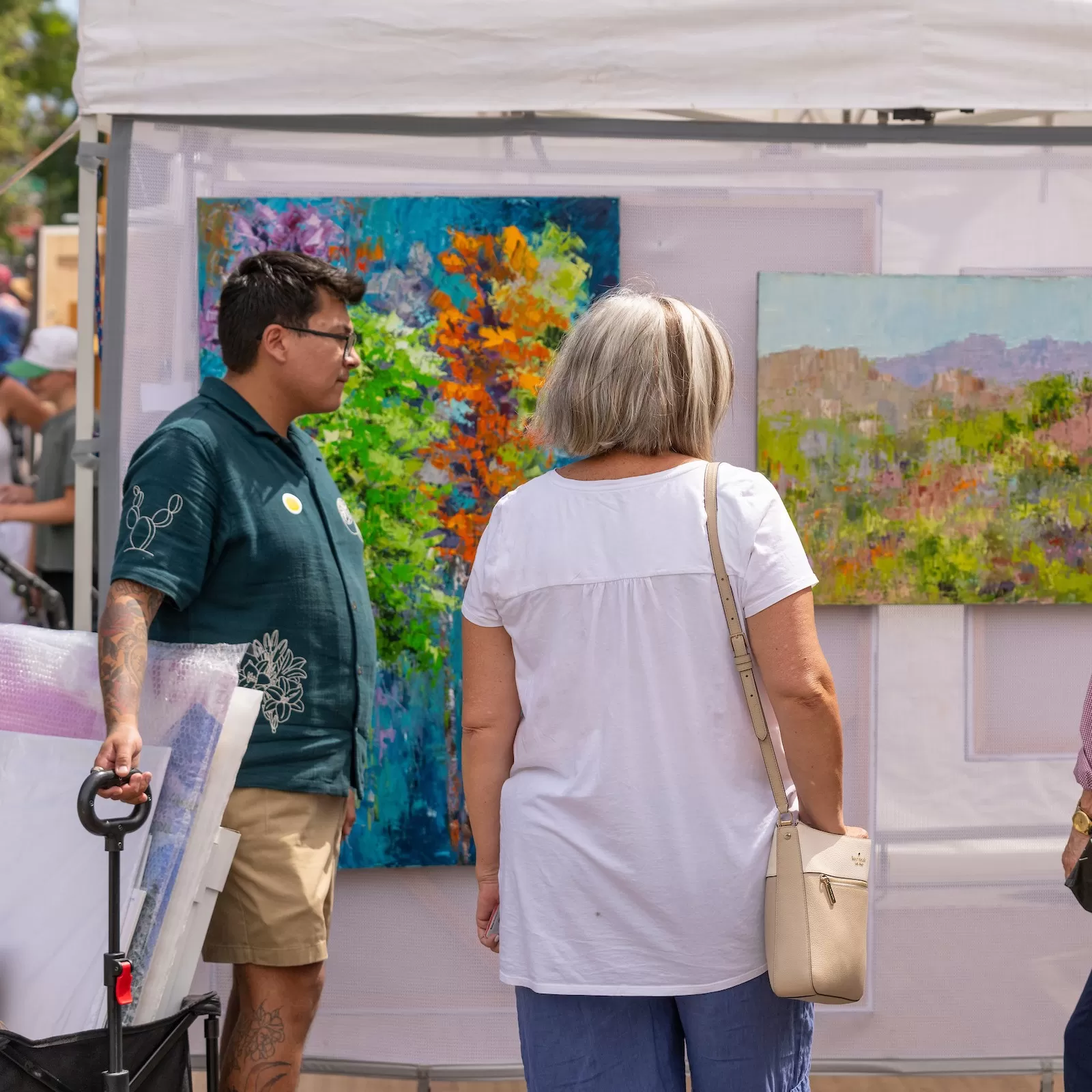 A visitor with a black wagon with artwork inside and another visitor with a purse looking at paintings on the outside of an artist tent booth.